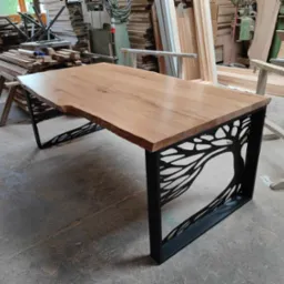 Stół z blatem z litego drewna o nieregularnych krawędziach i metalowych nogach z wyciętym motywem drzewa, widok z perspektywy podłogi w warsztacie stolarskim.