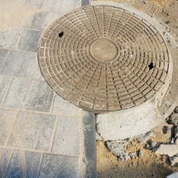 ART GREEN ARTEM ZADOROZHNII - Szara kostka brukowa ułożona wokół okrągłej studzienki kanalizacyjnej, widoczny but roboczy i szczotka do zamiatania, w tle ułożone rusztowanie.