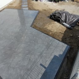 ART GREEN ARTEM ZADOROZHNII - Wykończony taras z szarych płyt z ciemną obwódką, widok z góry. Część terenu przygotowana pod dalsze prace, przykryta folią. Widoczny fragment ogrodzenia.