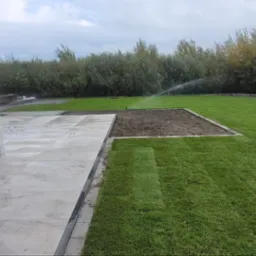 Nowoczesny taras z jasnych płyt, obrzeża z kostki, trawnik z nawadnianiem. Widok na ogród z drzewami w tle, pochmurne niebo.