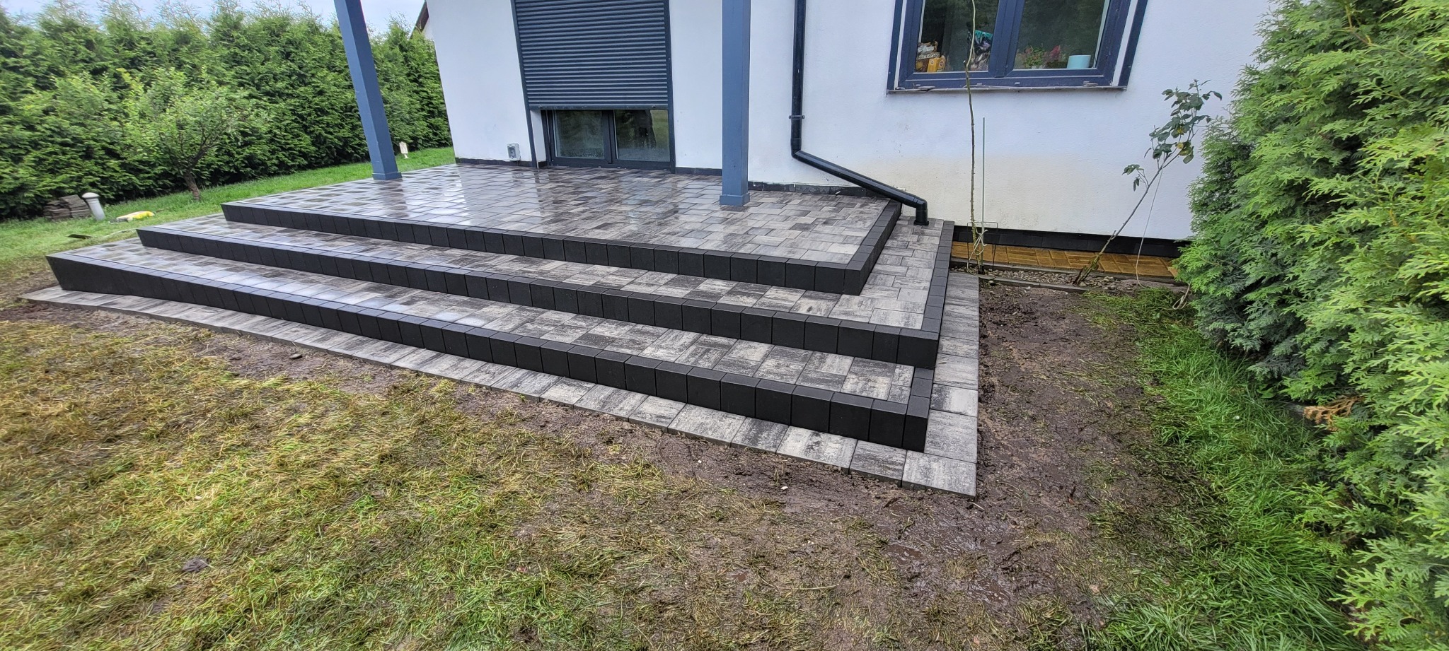 Schody z kostki brukowej prowadzące do domu, z czarnym obramowaniem, widoczne z poziomu trawnika. Elementy kostki ułożone w szachownicę.