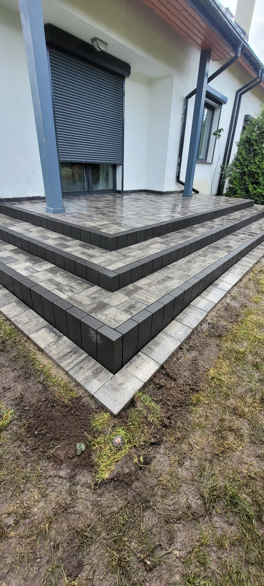 Nowoczesne schody wejściowe z kostki brukowej w odcieniach szarości, prowadzące do domu z szarą roletą okienną. Widok z dołu, na pierwszym planie trawa z ziemią.