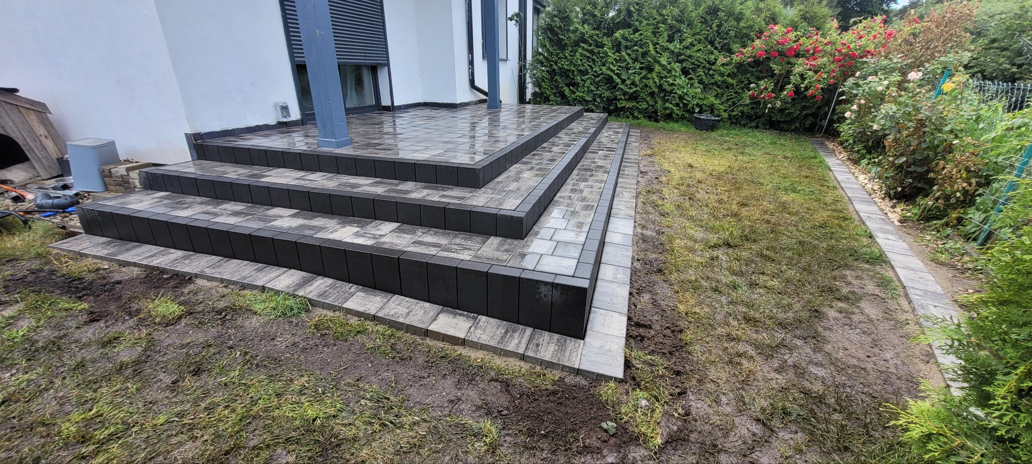 Zewnętrzne schody z kostki brukowej prowadzące do domu, z widocznym fragmentem ścieżki w ogrodzie. Kostka w odcieniach szarości i czerni, ułożona w geometryczny wzór.