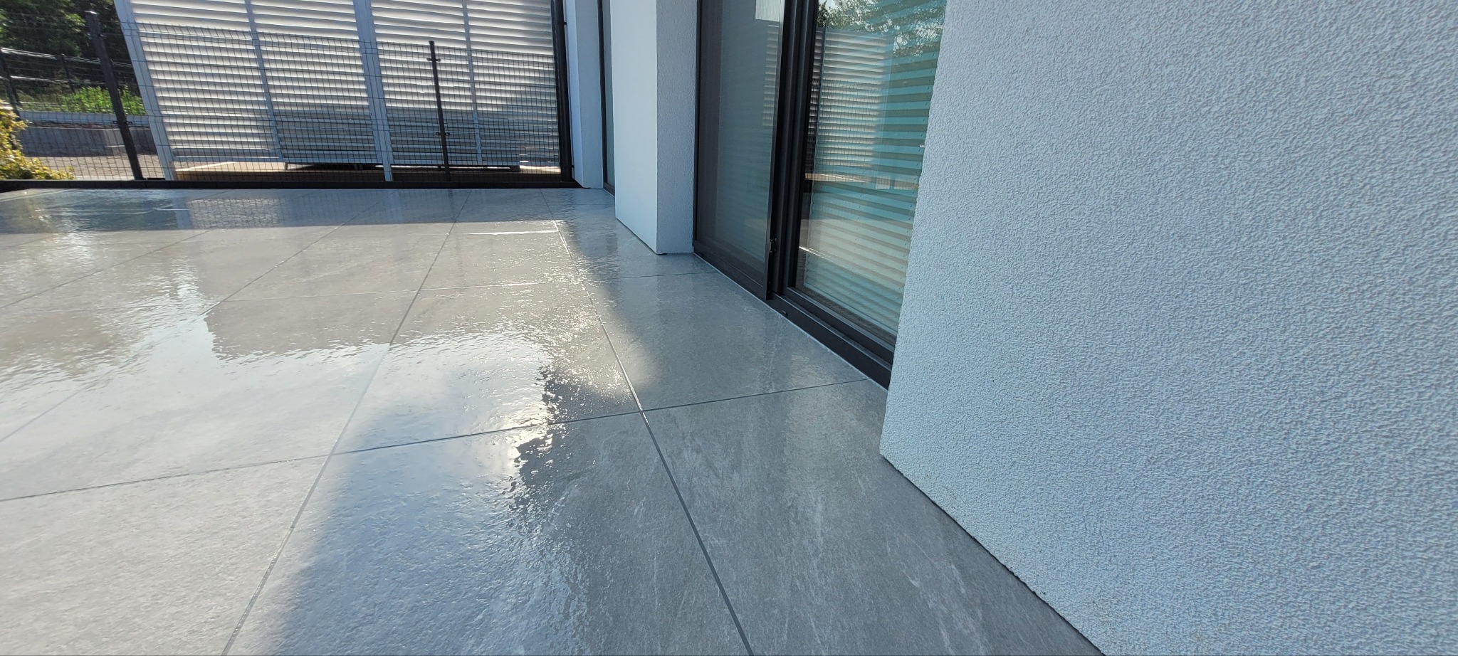 Błyszcząca, szara, ceramiczna podłoga tarasu przylegająca do ściany budynku z oknem przesuwnym. Widoczne odbicia światła na powierzchni płytek.