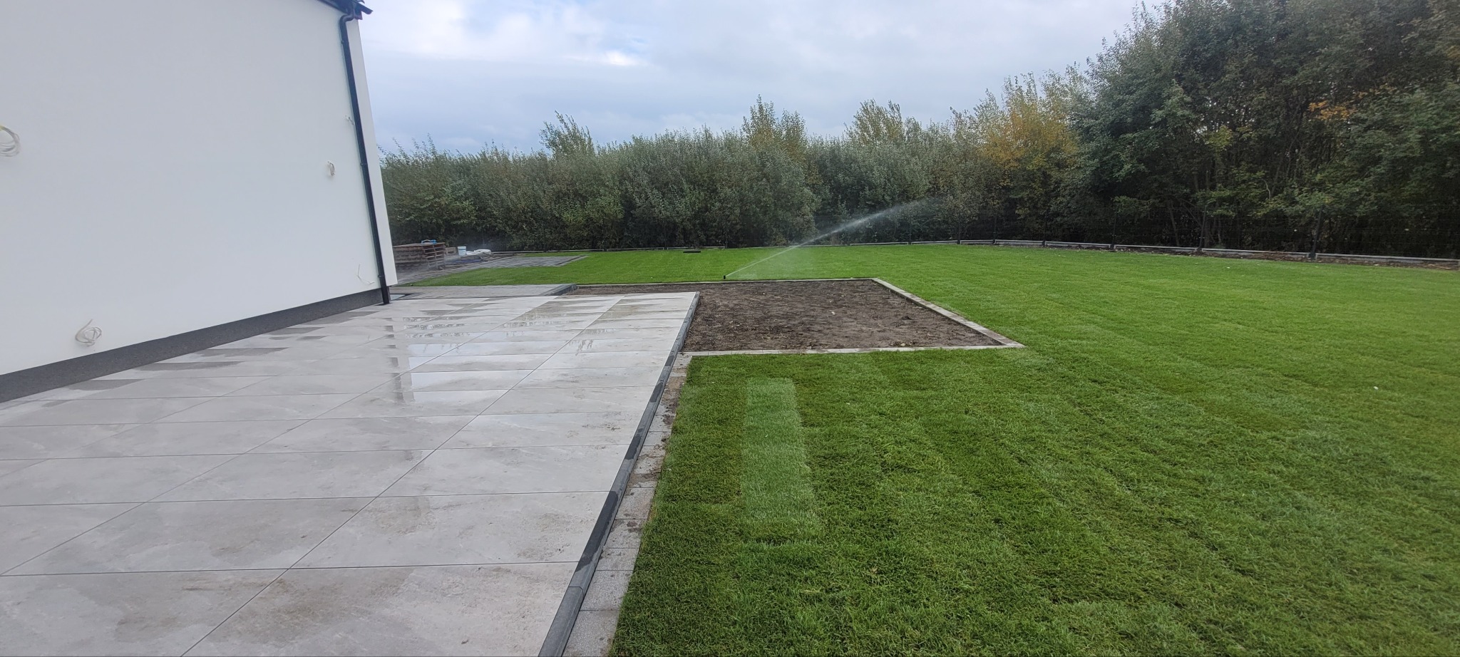 Nowoczesny taras z jasnych płyt, obrzeża z kostki, trawnik z nawadnianiem. Widok na ogród z drzewami w tle, pochmurne niebo.
