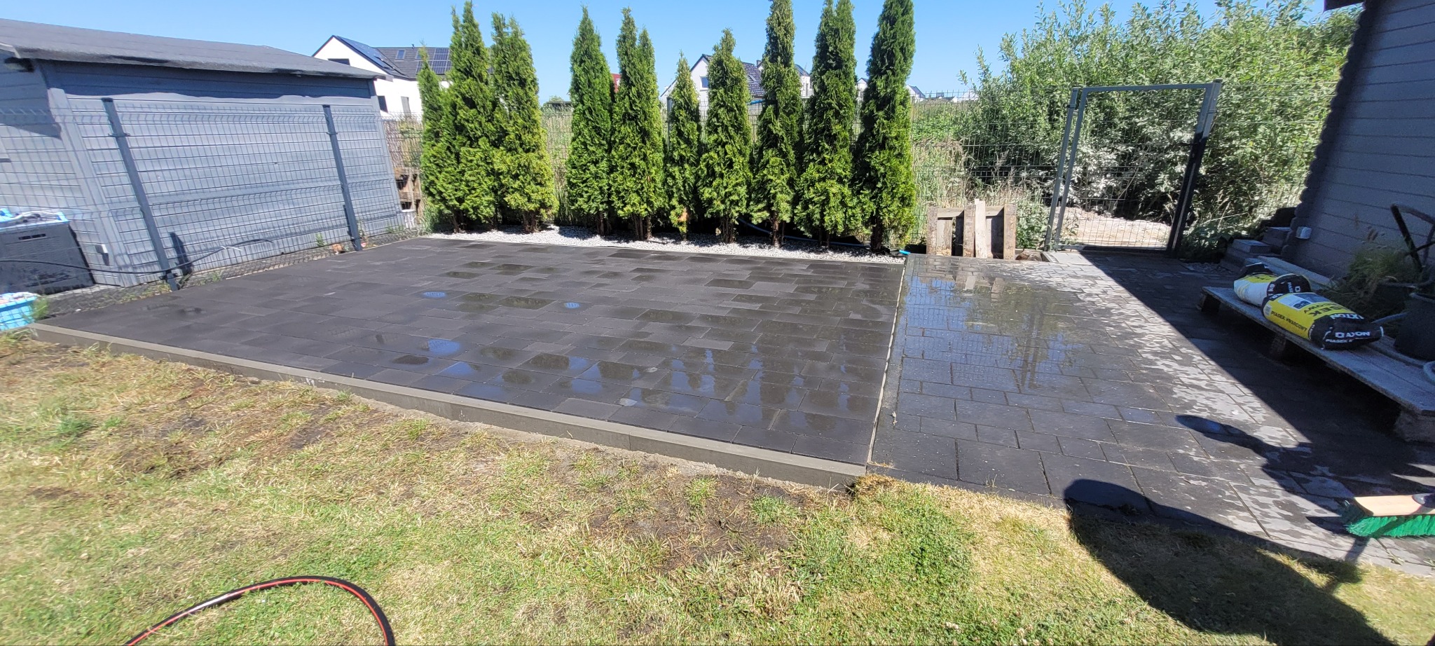 Ułożona, ciemnoszara kostka brukowa na posesji, obok zielone tuje i trawnik. Część kostki jest mokra, odbija światło. W tle widoczny szary budynek gospodarczy.