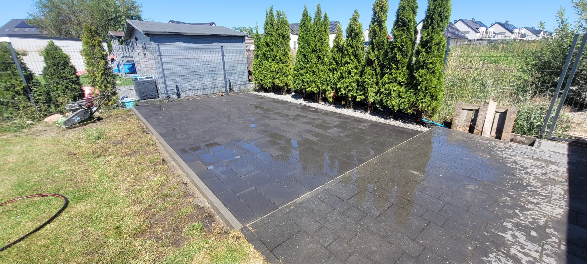 Ciemnoszara kostka brukowa ułożona na posesji, częściowo mokra, z widoczną ubijarką w tle i tujami wzdłuż ogrodzenia. Słoneczny dzień.