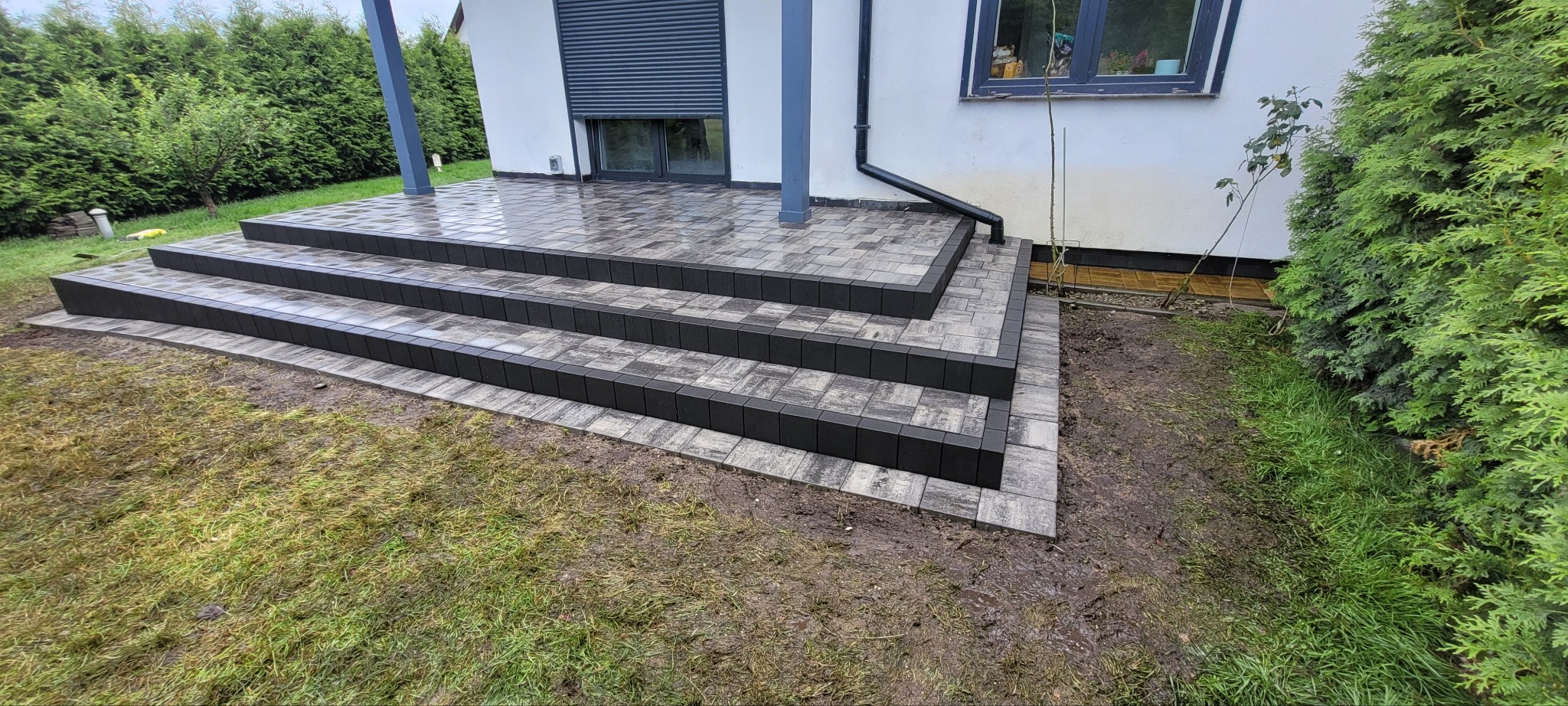 Schody z kostki brukowej prowadzące do domu z szarą elewacją i zielonymi roślinami w tle. Kostka ułożona w geometryczny wzór, widoczne detale wykończenia.