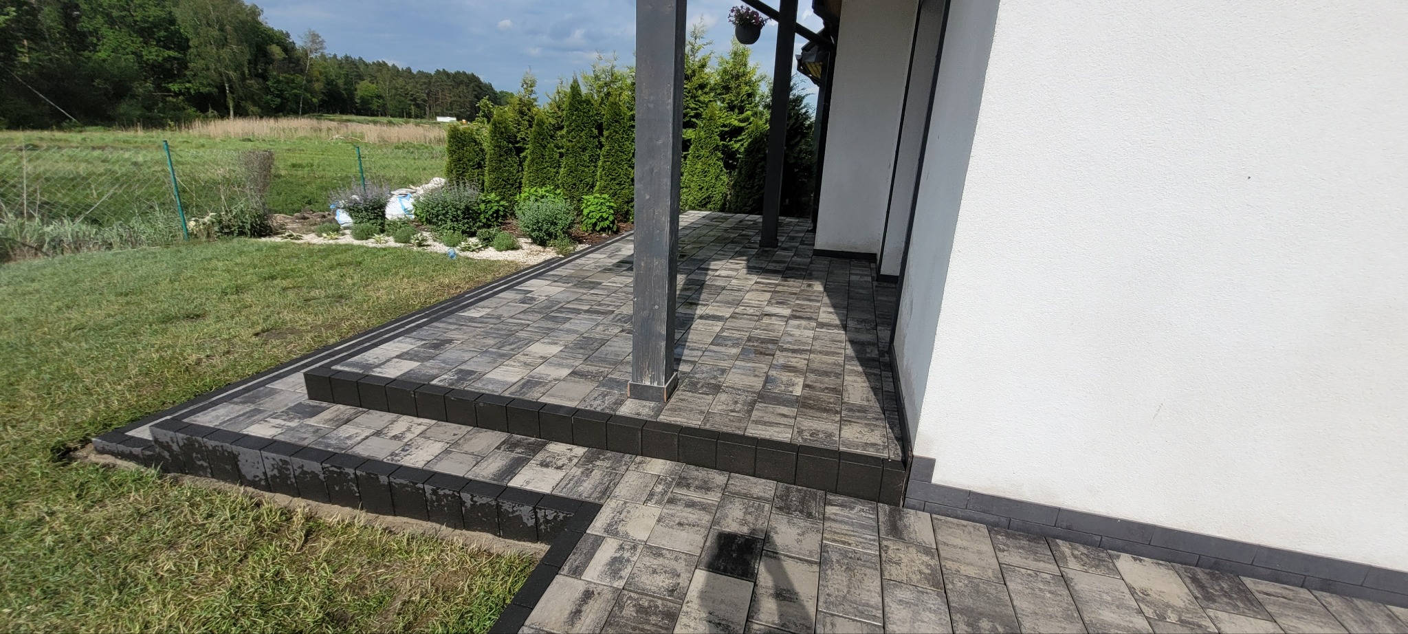 Ułożona kostka brukowa w odcieniach szarości tworząca ścieżkę i schody przy budynku z ogrodem w tle. Widoczny fragment podpory z ciemnego drewna.