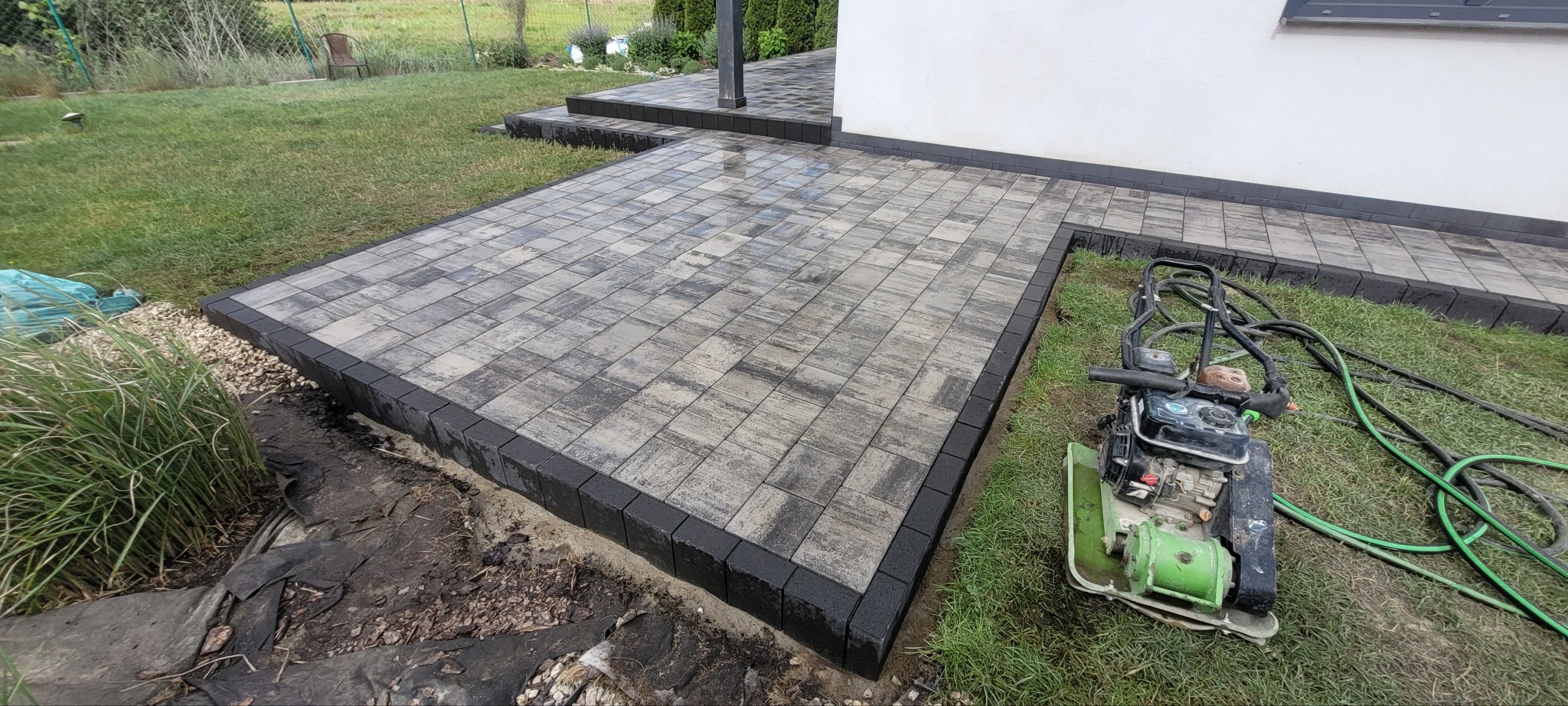Świeżo ułożona, szara kostka brukowa z czarnym obramowaniem, prowadząca do domu. Obok stoi zagęszczarka w kolorze zielonym. Widok na trawnik i fragment budynku.