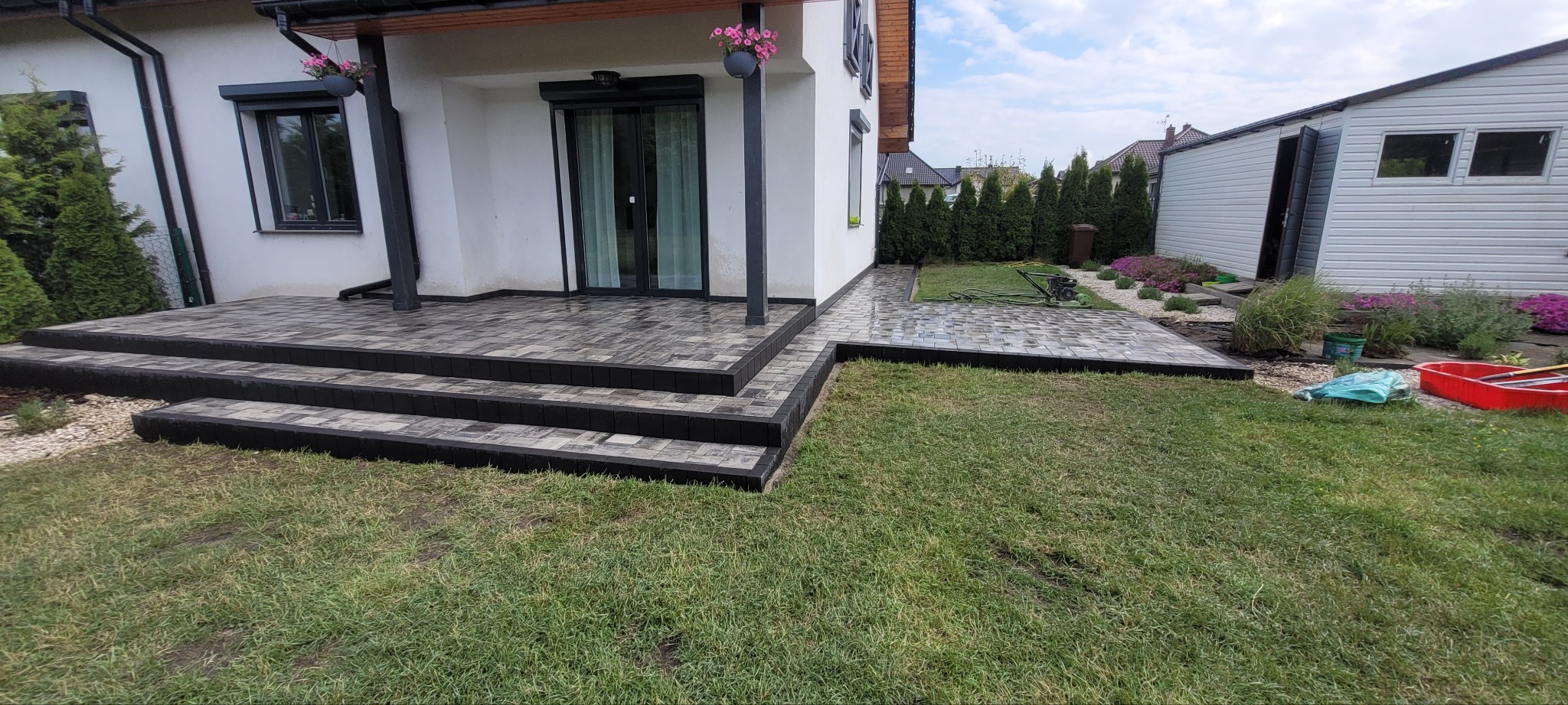 Ułożona kostka brukowa w odcieniach szarości tworzy schody i ścieżkę przy domu z białymi ścianami. Widoczny trawnik, rośliny ozdobne i budynek gospodarczy w tle.