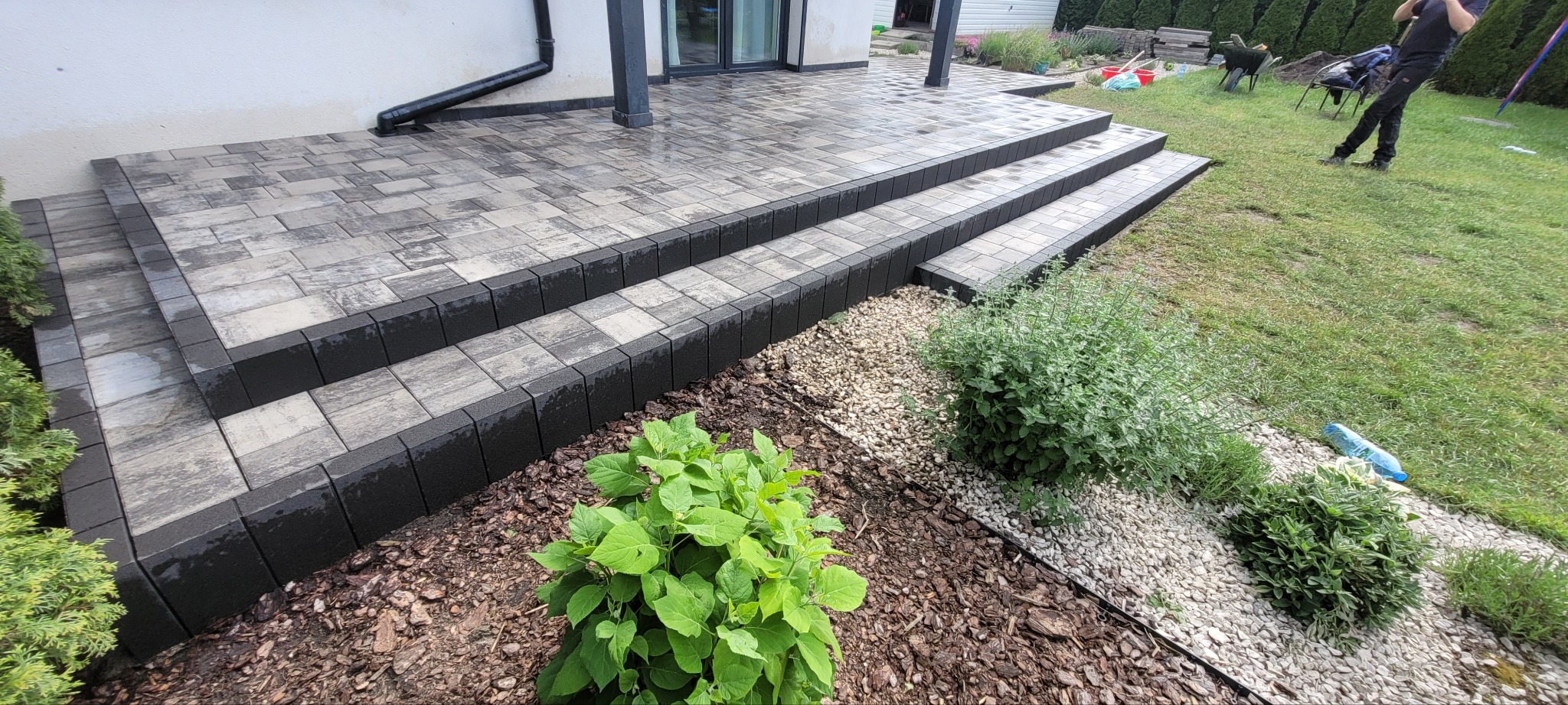 Schody i taras z kostki brukowej w odcieniach szarości, obrzeża w kolorze czarnym. Widoczne rośliny ozdobne i trawnik. Nowoczesne wykończenie przestrzeni przydomowej.