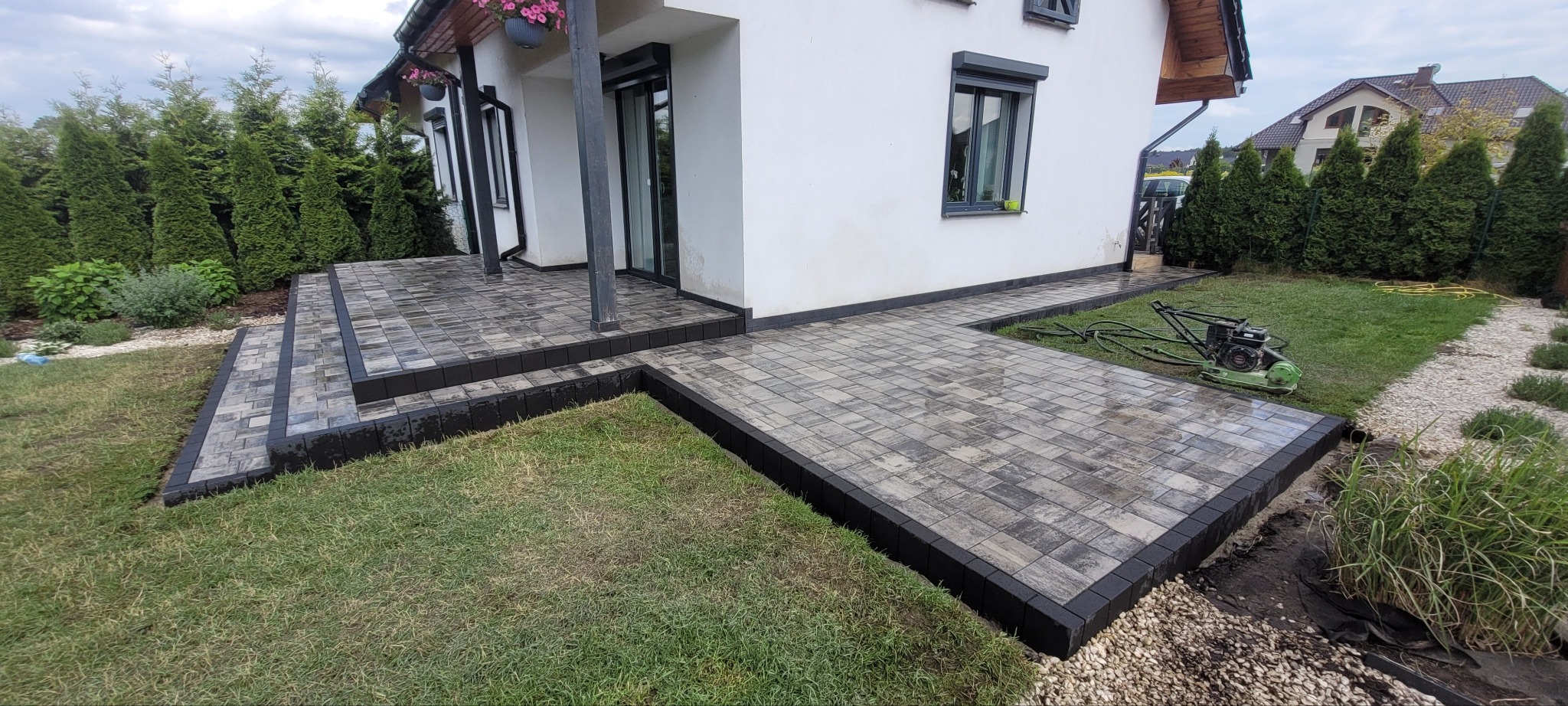 Nowoczesny taras i ścieżka z kostki brukowej w odcieniach szarości, obrzeża z ciemnej kostki. W tle biały dom z ciemnymi oknami i zielony trawnik.
