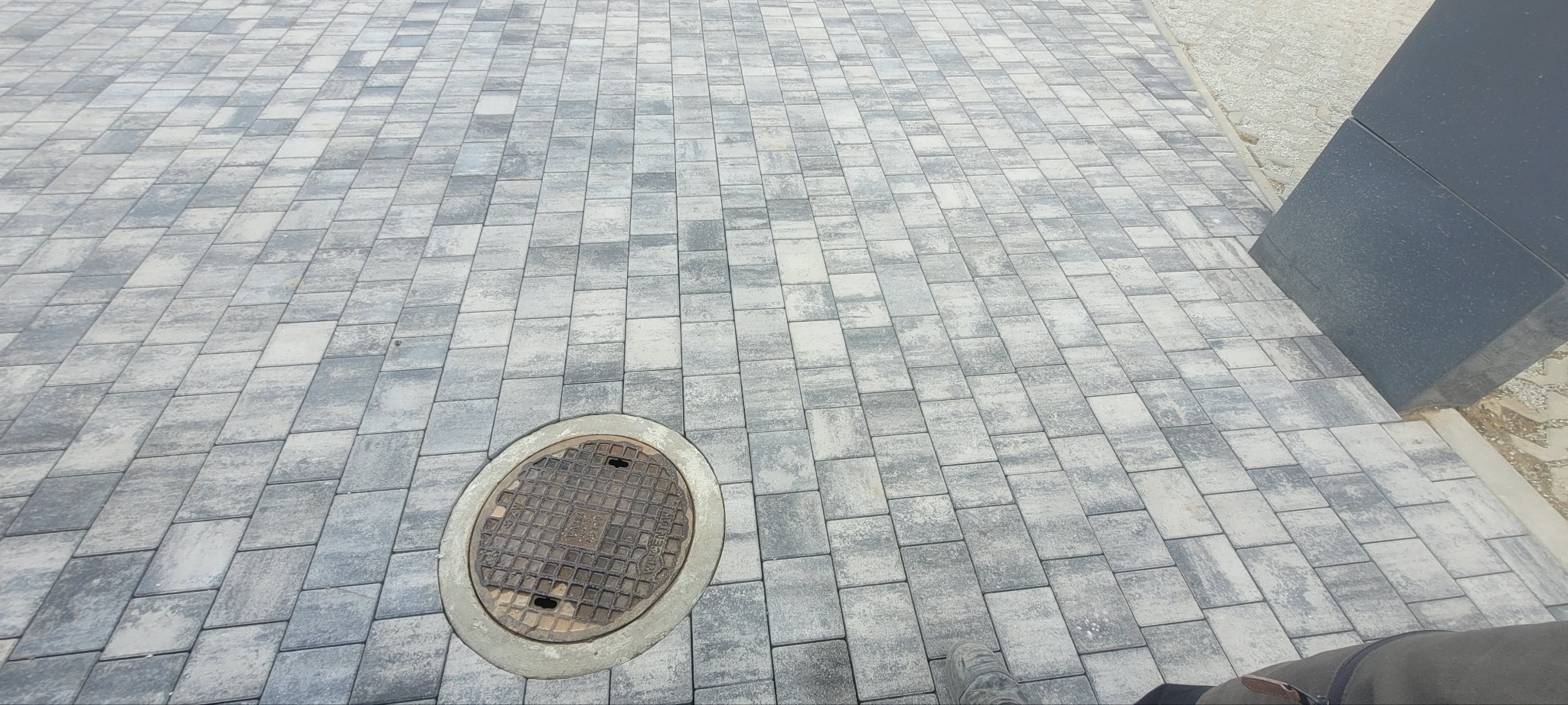 Szara kostka brukowa ułożona wokół studzienki kanalizacyjnej, widok z góry, fragment buta w rogu kadru, nowoczesna architektura w tle.