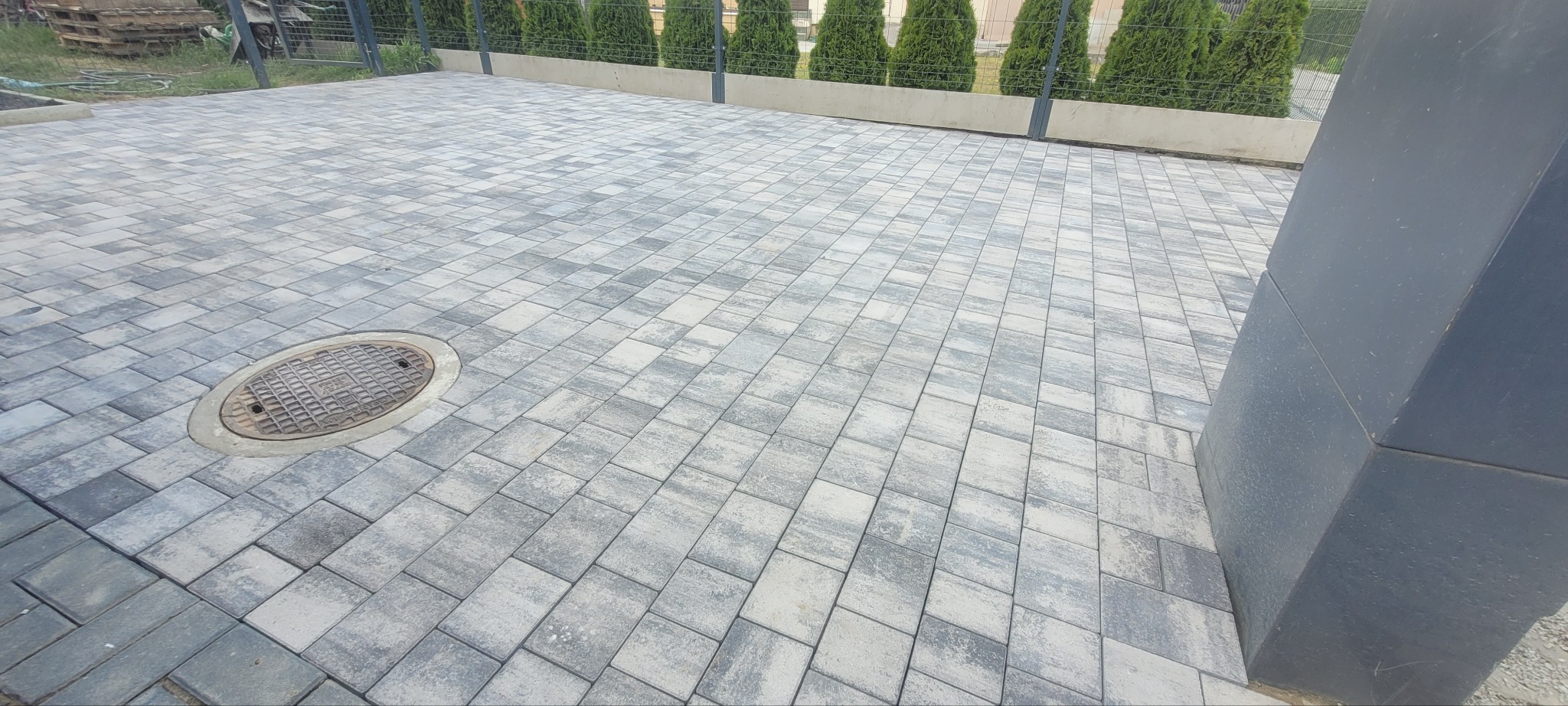 Szara kostka brukowa ułożona w jodełkę, z widoczną studzienką kanalizacyjną i rzędem tuj w tle. Ujęcie z niskiej perspektywy, podkreślające wzór i fakturę kostki.