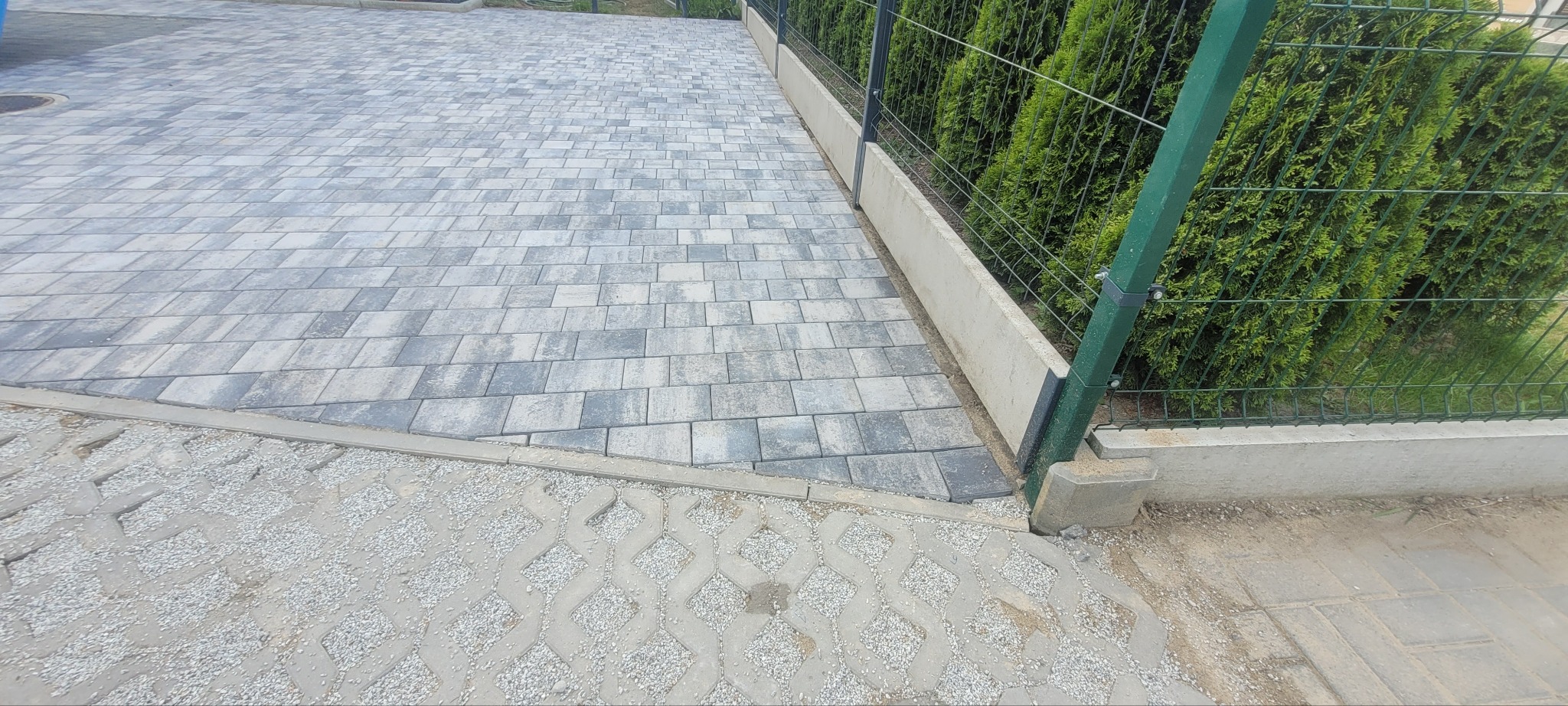 Szara kostka brukowa ułożona na podjeździe, obok ażurowa kostka z wypełnieniem żwirowym i zielony płot z tujami w tle. Widok z góry, lekki kąt.