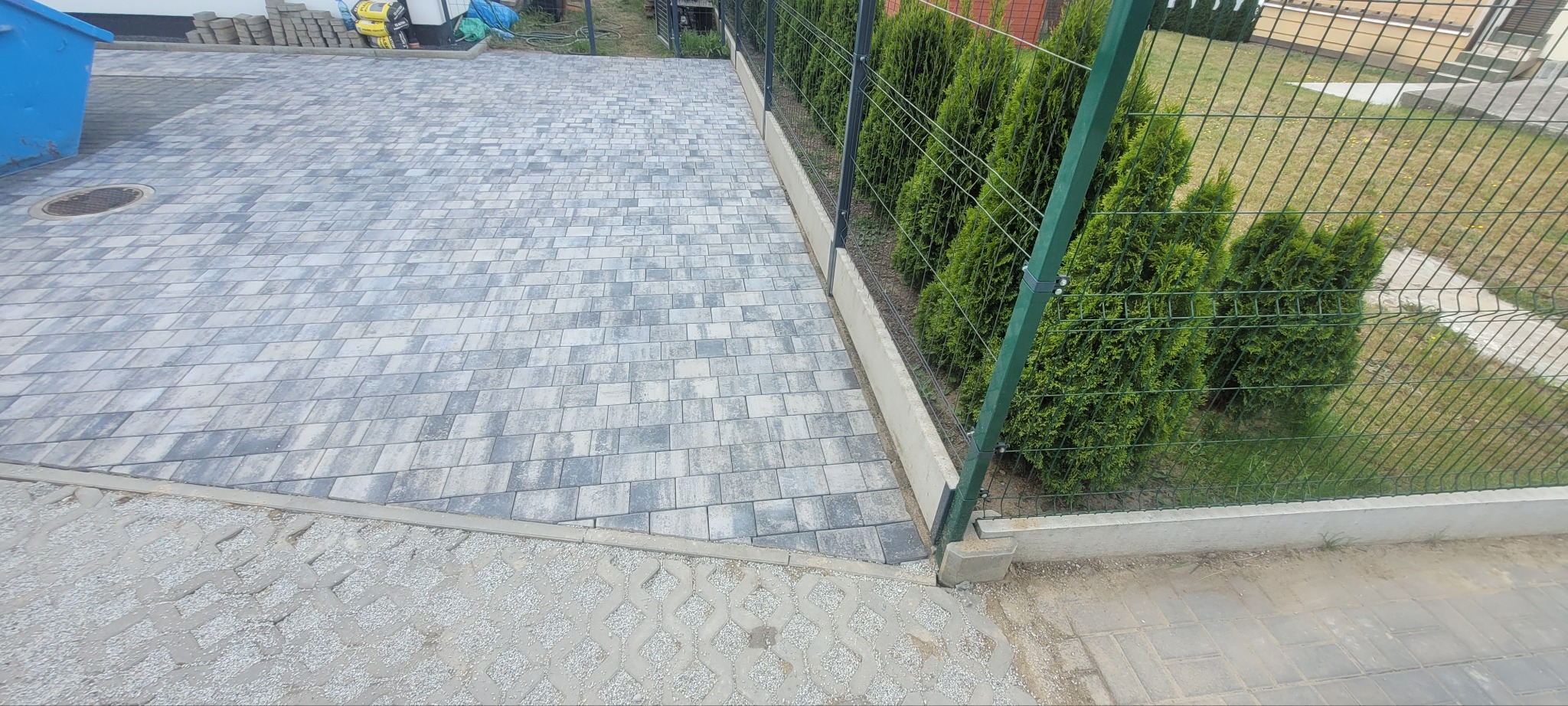Szara kostka brukowa ułożona wzorem jodełki, obrzeża betonowe, siatka ogrodzeniowa z zielonymi tujami w tle. Widok z góry na gotową nawierzchnię.