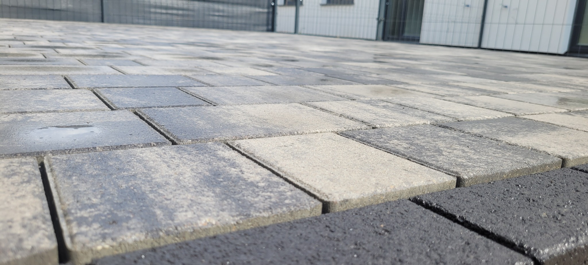 Ułożona, wielobarwna kostka brukowa w odcieniach szarości i beżu, z widocznymi kroplami wody, tworząca równą powierzchnię przed budynkiem.