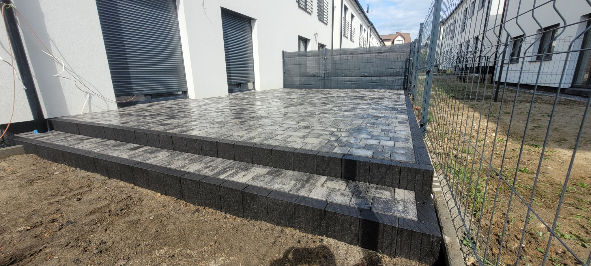 Ułożona kostka brukowa w szarościach tworząca schody i taras przy budynku z szarą siatką ogrodzeniową w tle. Widok z boku.