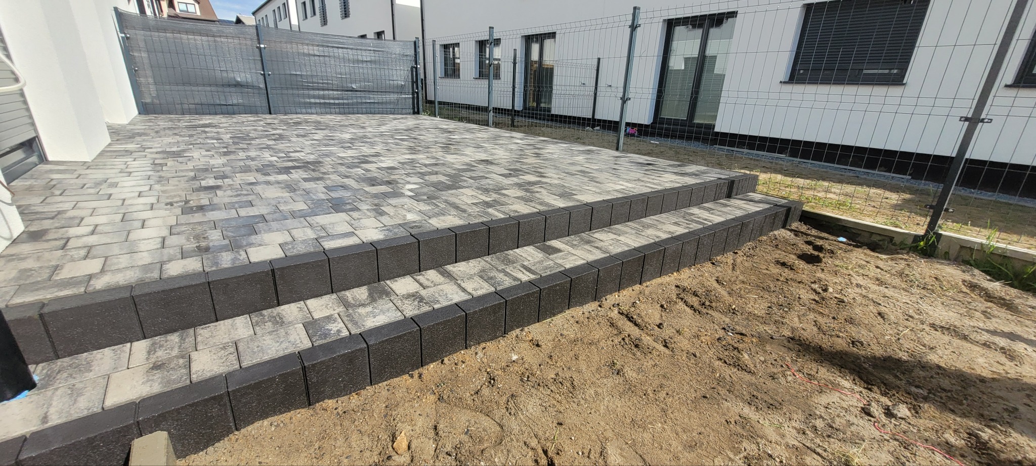 Ułożona kostka brukowa w Szczecinie, tworząca taras z dwoma stopniami. Kostka szaro-beżowa, obrzeża czarne. Widoczne ogrodzenie i budynek w tle.