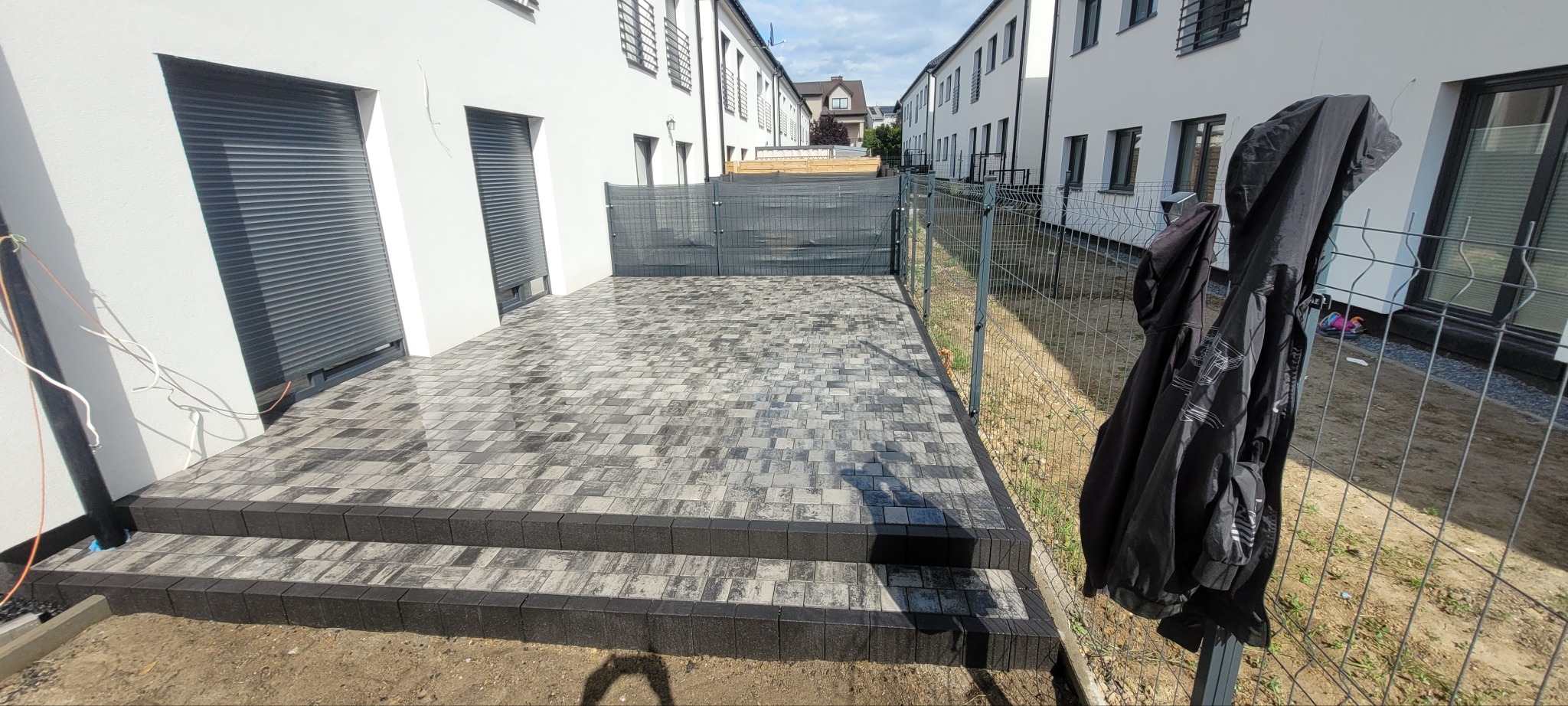 Ułożona kostka brukowa w odcieniach szarości prowadząca do domu z szarą bramą. Widoczne dwa stopnie i fragment ogrodzenia z powieszoną kurtką.