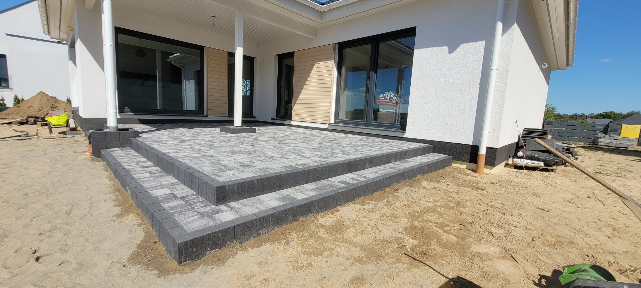 Wykończony taras z kostki brukowej w odcieniach szarości, z niskimi schodami, przylegający do białego domu z dużymi oknami. Widoczne pozostałości piasku po pracach.
