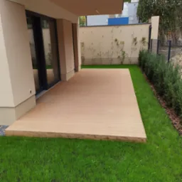 Jasny taras z desek kompozytowych przylegający do budynku, otoczony świeżą zieloną trawą i niskimi krzewami, z widocznym fragmentem elewacji budynku i ogrodzenia w tle.