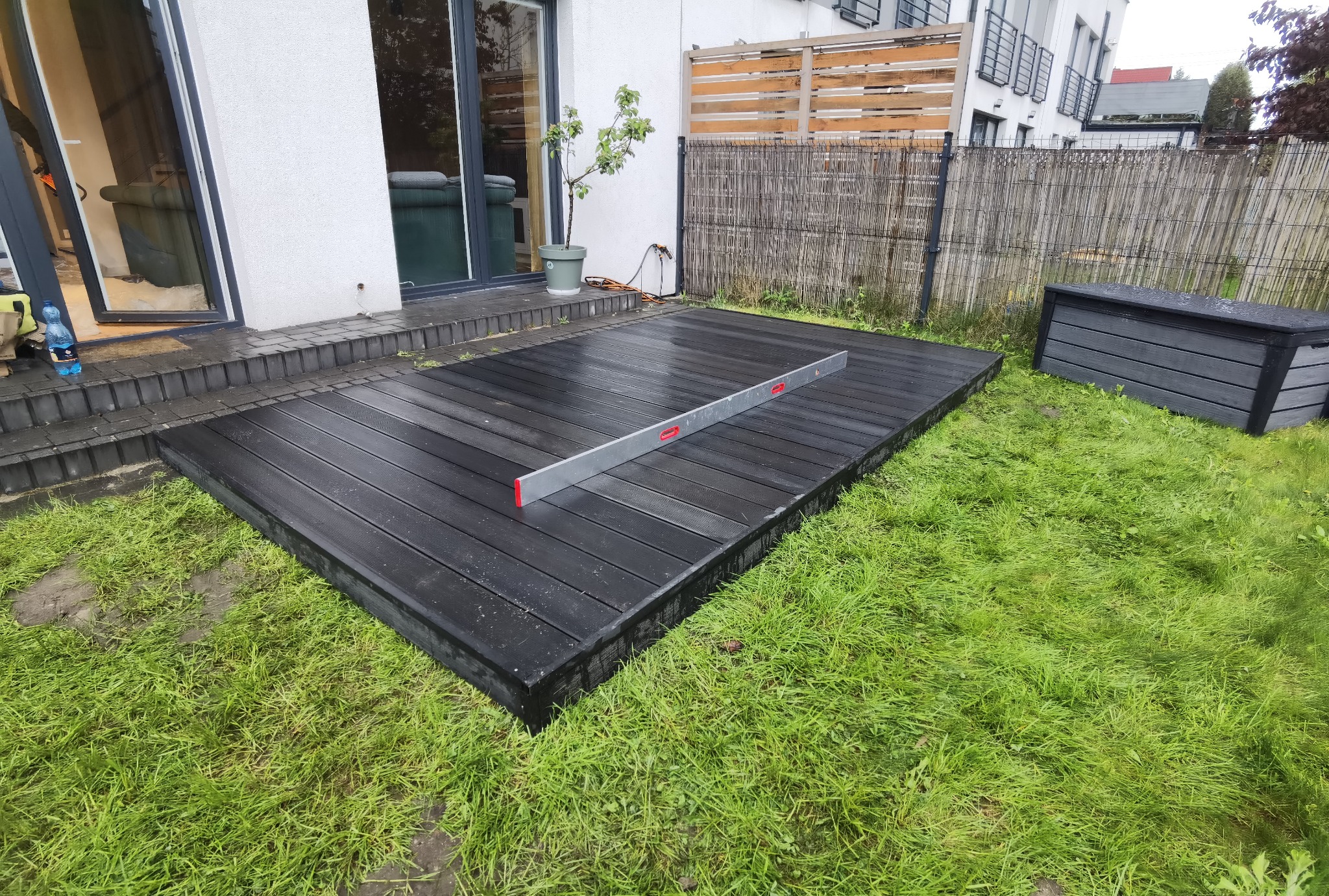 Montaż czarnego tarasu z desek kompozytowych, poziomowany za pomocą poziomicy, obok skrzynia ogrodowa z tego samego materiału, widoczny trawnik i fragment domu.