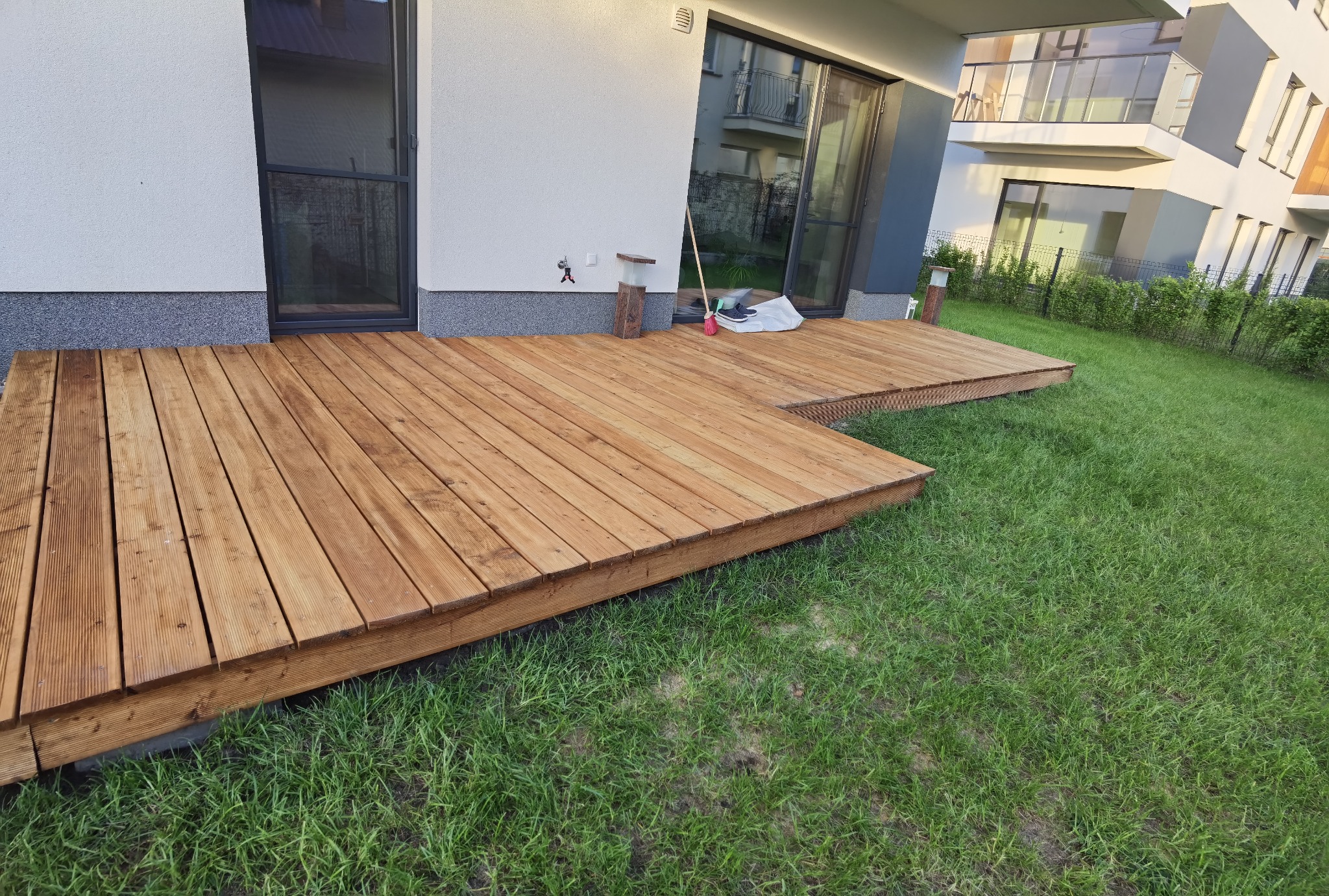 Nowoczesny taras z drewna, przylegający do budynku z dużymi oknami, z widoczną szczotką i butami na krawędzi tarasu.