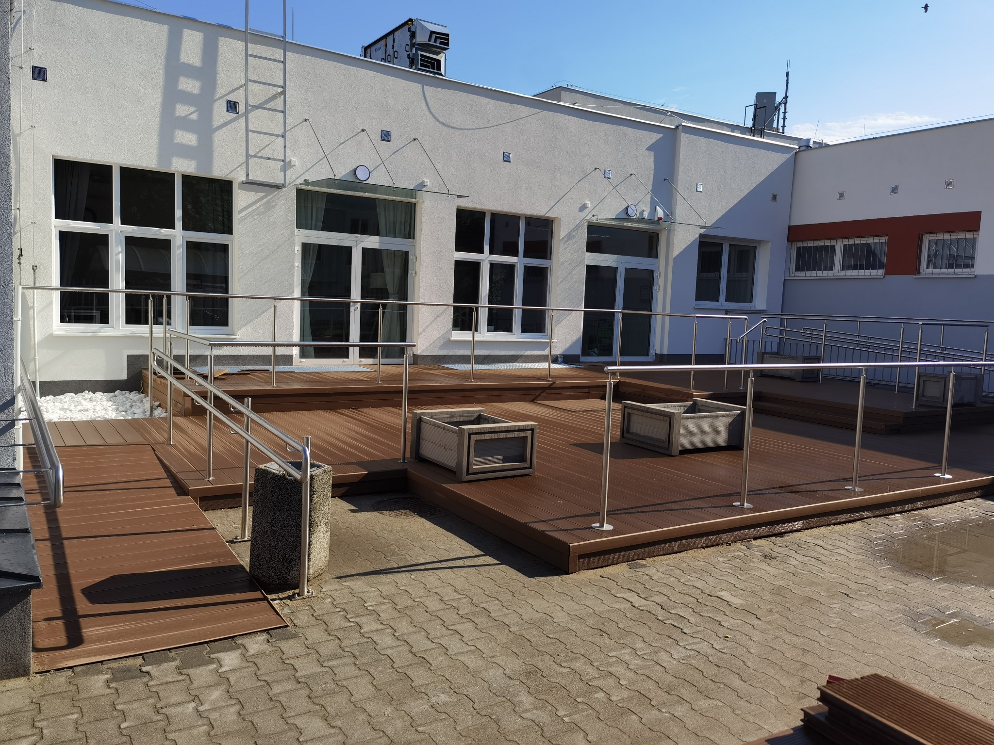 Zewnętrzny taras z desek kompozytowych z metalową balustradą, schodami i donicami, przylegający do budynku z białymi oknami i drabiną pożarową.