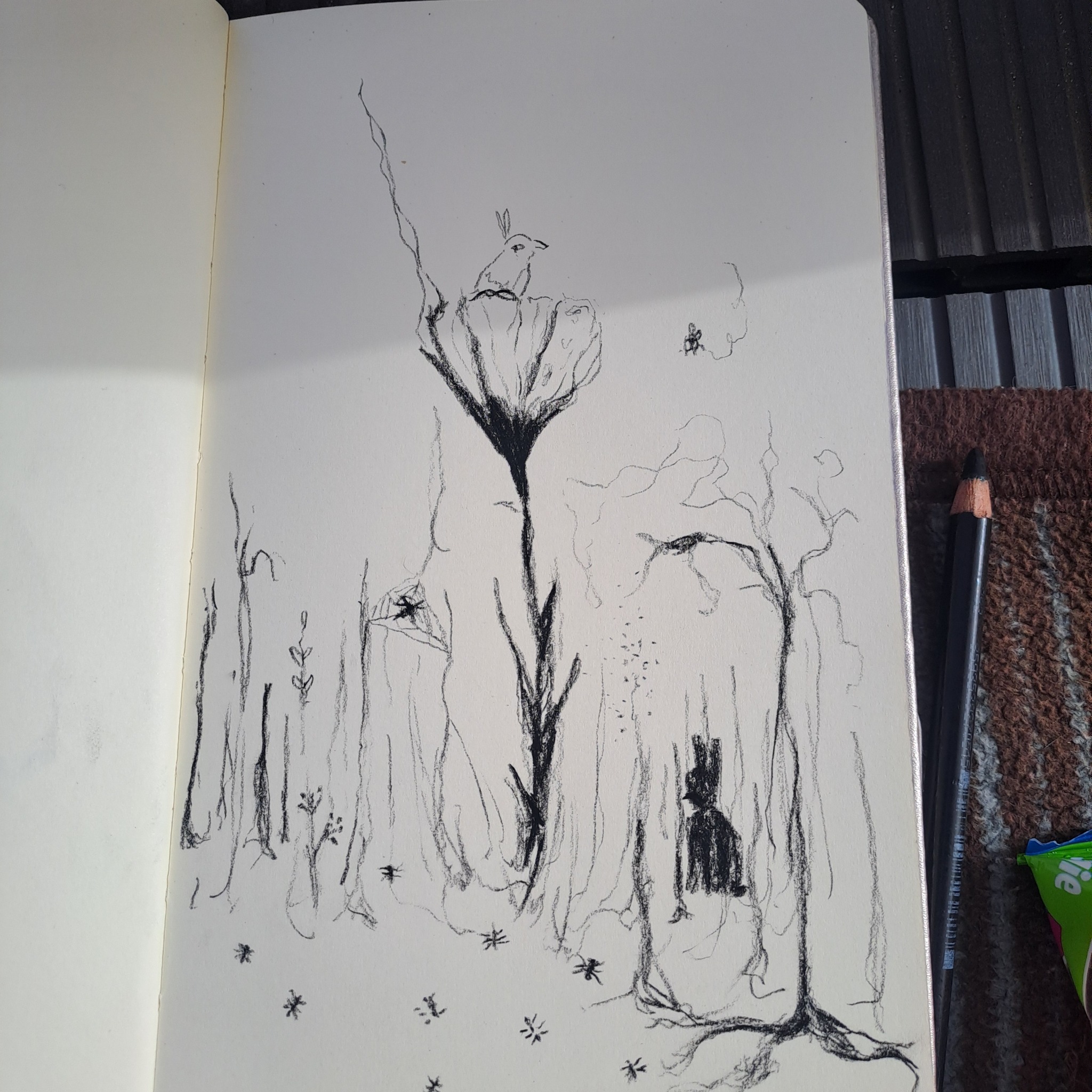 Szkic w czerni przedstawiający fantazyjny krajobraz z motywem królika na kwiatku, wykonany ołówkiem w otwartym szkicowniku leżącym na brązowej powierzchni.