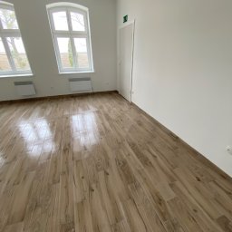 AJP INVESTED Adam Porębny - Wnętrze pokoju po remoncie, widoczne panele podłogowe imitujące drewno, białe ściany, dwa okna z widokiem na drzewa oraz białe grzejniki.