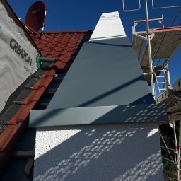 AJP INVESTED Adam Porębny - Fragment elewacji budynku z widocznym ociepleniem, częściowo pokrytym szarą blachą, oraz rusztowaniem i anteną satelitarną na dachu z czerwonej dachówki.