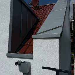 AJP INVESTED Adam Porębny - Detale wykończenia dachu i elewacji budynku z widoczną rynną, obróbką blacharską komina i anteną satelitarną na tle błękitnego nieba z chmurami.