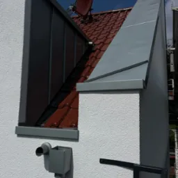 Detale wykończenia dachu i elewacji budynku z widoczną rynną, obróbką blacharską komina i anteną satelitarną na tle błękitnego nieba z chmurami.