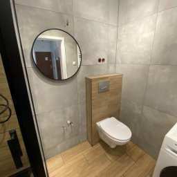 AJP INVESTED Adam Porębny - Nowoczesna łazienka w szarościach z drewnopodobną podłogą i okrągłym lustrem, widoczne elementy instalacji przed wykończeniem.