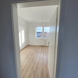 AJP INVESTED Adam Porębny - Pusty pokój po remoncie, widok z korytarza przez otwarte drzwi, jasne panele podłogowe, białe ściany, okno z widokiem na blok i drzewa bez liści, biały grzejnik pod oknem, wiszące kable...