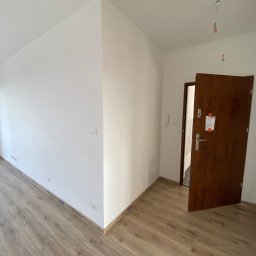 AJP INVESTED Adam Porębny - Puste, świeżo wykończone wnętrze mieszkania z panelami podłogowymi i białymi ścianami, brązowe drzwi wejściowe uchylone, widoczne gniazdka elektryczne i wystające przewody na suficie.