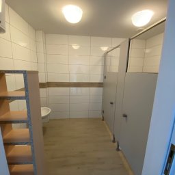 AJP INVESTED Adam Porębny - Wnętrze publicznej toalety z kabinami, białe płytki na ścianach, jasna podłoga, widoczny fragment niedokończonego regału z płyty wiórowej.