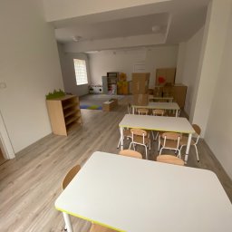 AJP INVESTED Adam Porębny - Jasne, przestronne wnętrze sali przedszkolnej z białymi stolikami, drewnianymi krzesłami i laminowaną podłogą imitującą drewno, z widocznym kącikiem zabaw i regałami w tle.
