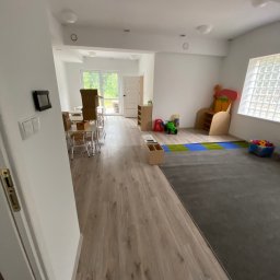 AJP INVESTED Adam Porębny - Przestronne, nowocześnie wykończone wnętrze sali zabaw z drewnianą podłogą, szarą wykładziną dywanową, białymi ścianami i dużym oknem z luksferami; widoczne zabawki, stoliki z krzesłami i kartonowe...