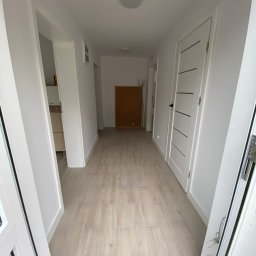 AJP INVESTED Adam Porębny - Jasny korytarz z białymi ścianami, panelami podłogowymi imitującymi drewno i białymi drzwiami z poziomymi wgłębieniami, widziany z perspektywy wejścia, z widocznym w tle wieszakiem na ubrania.