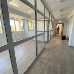 AJP INVESTED Adam Porębny - Korytarz biurowy z przeszklonymi ściankami działowymi i podłogą imitującą drewno; w tle widoczna osoba w pomarańczowej koszulce.