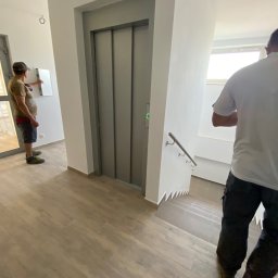 AJP INVESTED Adam Porębny - Wnętrze klatki schodowej z windą, pracownik sprawdza skrzynkę elektryczną, widoczne drewnopodobne panele podłogowe i schody wyłożone płytkami.