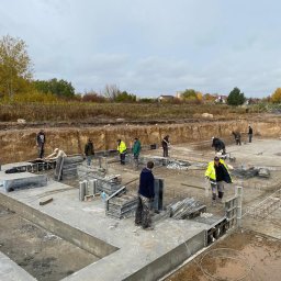 AJP INVESTED Adam Porębny - Budowa fundamentów: ekipa budowlana pracuje nad wylewaniem betonu, widoczne szalunki, zbrojenia i wykop pod budynek, w tle krajobraz z zabudowaniami.