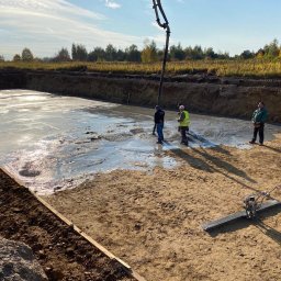 AJP INVESTED Adam Porębny - Wylewanie fundamentów pod budynek w Grudziądzu, widoczna pompa do betonu, pracownicy w kaskach i kamizelkach odblaskowych oraz zacieraczka do betonu.