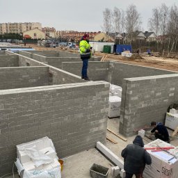 AJP INVESTED Adam Porębny - Budowa domu z bloczków silikatowych: widok na fundamenty z murami, pracownicy w odzieży roboczej, palety z bloczkami, otoczenie budowy z budynkami mieszkalnymi w tle.