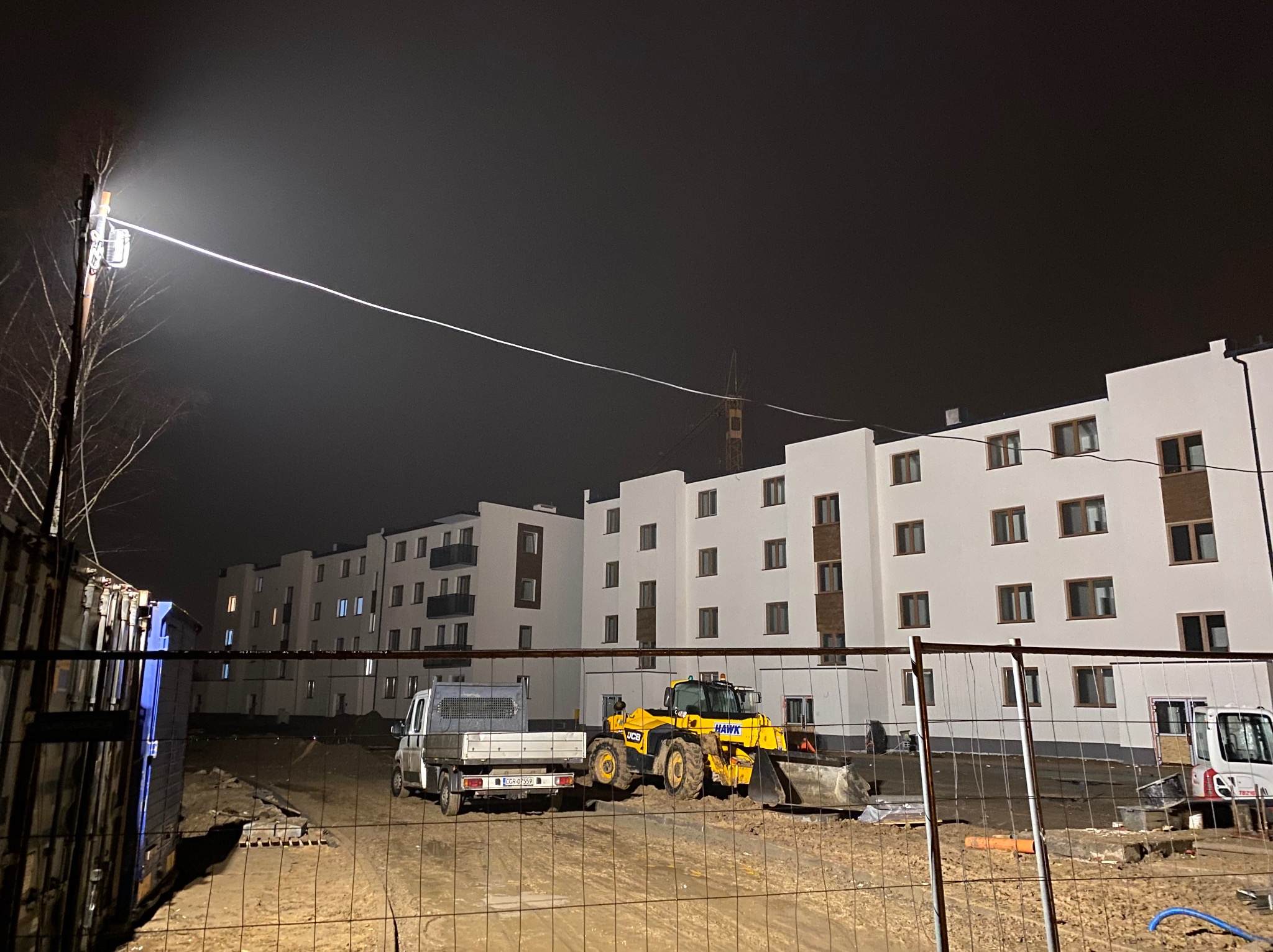 Nowe, ocieplone bloki mieszkalne w trakcie budowy, widoczne maszyny budowlane: ładowarka JCB i samochód dostawczy, oświetlone przez reflektor budowlany w nocy.