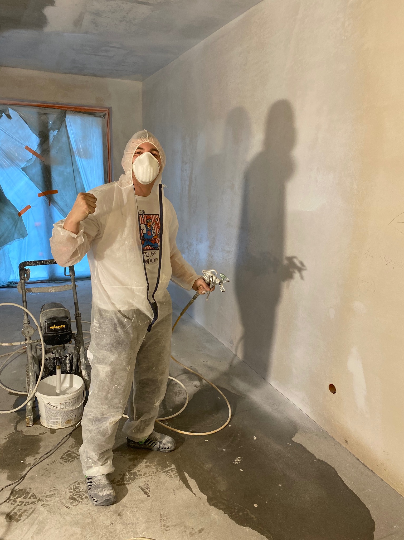 Pracownik w kombinezonie ochronnym i masce, z pistoletem natryskowym, prezentuje świeżo pomalowaną ścianę w pomieszczeniu w trakcie wykańczania, z widocznym agregatem malarskim i śladami farby...