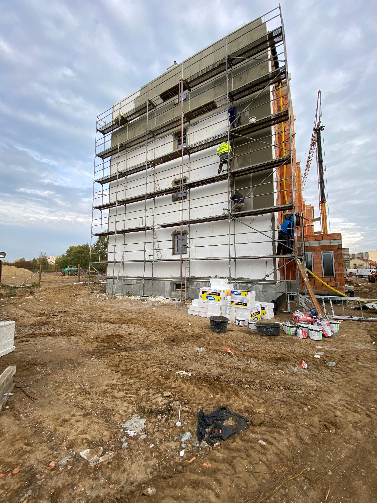 Prace ociepleniowe na elewacji budynku w trakcie realizacji, widoczne rusztowania, styropian i pracownicy w odzieży roboczej, obok budynek w trakcie budowy z czerwonej cegły, w tle dźwig...
